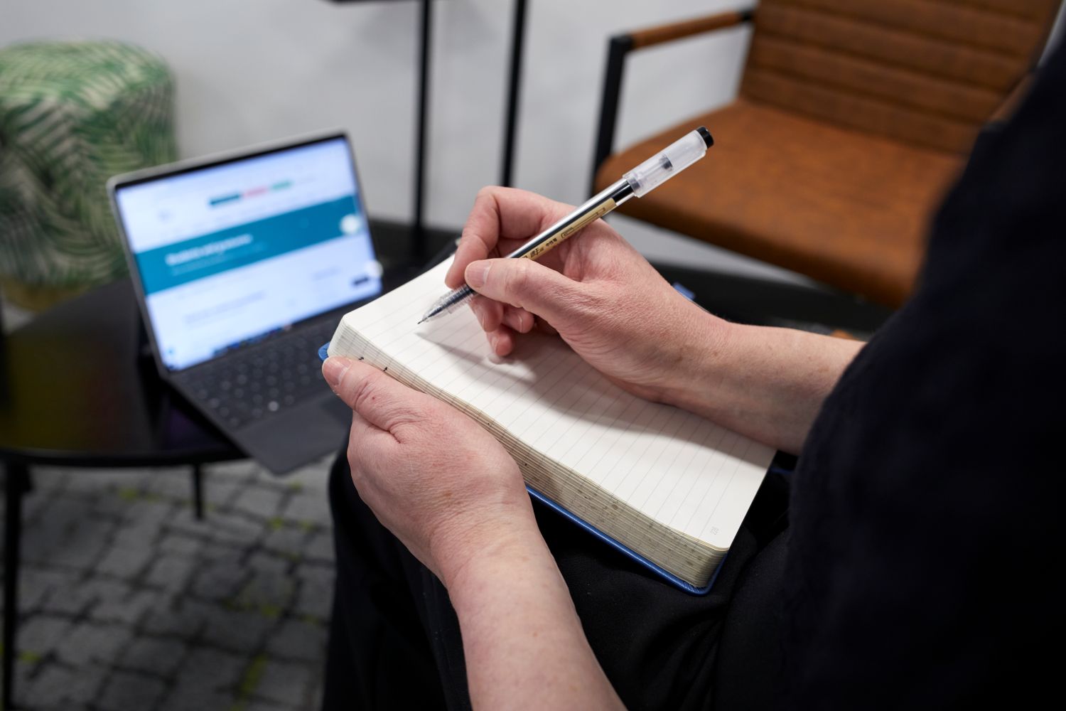 Someone writing in a notebook and holding a pen. A laptop is placed on a table in front of them.