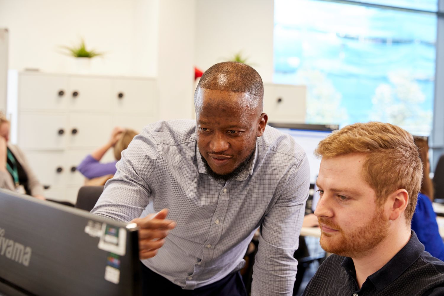 Two men looking at a computer monitor. One is sat down at a chair, and another is stood up and pointing at the screen.